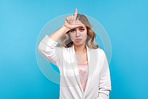 I`m loser. Portrait of depressed woman doing loser gesture, L finger sign on forehead. blue background