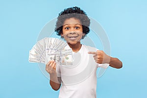 I`m rich! Joyful lucky wealthy little boy with curly hair pointing to fan of dollar banknotes