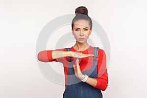 I need break! Portrait of tired displeased girl with hair bun in denim overalls showing time out gesture and looking dissatisfied