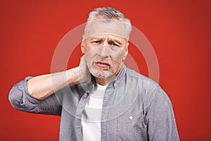 I need a massage. Frustrated senior man in casual clothing holding hand on his neck while standing against red background