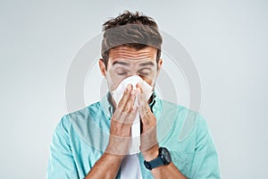 I need more tissues. Studio shot of a young man blowing his nose with a tissue while standing against a grey background.