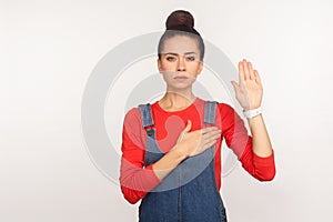 I promise! Portrait of trustworthy honest girl with hair bun in denim overalls raising palm to swear allegiance photo