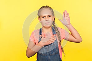 I promise to be honest! Portrait of serious responsible little girl with braid in denim overalls raising palm to take oath