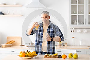 I recommend healthy food. Happy senior man drinking fresh juice and showing thumb up in kitchen interior