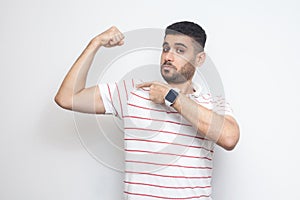 I am strong. Portrait of proud handsome bearded young man in striped t-shirt standing, pointing at his bicep and looking at camera