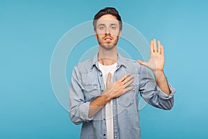 I swear! Portrait of honest responsible man in worker denim shirt giving evidence with raised hand, blue background