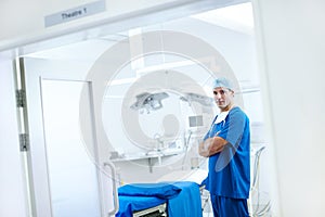 I take pride in my operating theatre. A handsome male doctor standing next to an operating table with his arms crossed.