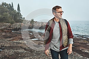 I walk this path everyday. a confident middle aged man walking on rocks next to the ocean while wearing a backpack