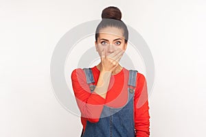 I will not say anything. Portrait of frightened intimidated girl with hair bun in denim overalls covering her mouth
