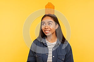 I wish own home! Portrait of happy girl in denim shirt standing with paper house on her head, dreaming of real estate