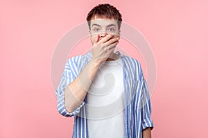 I won`t tell. Portrait of amazed scared brown-haired man in casual striped shirt covering mouth with hand. isolated on pink