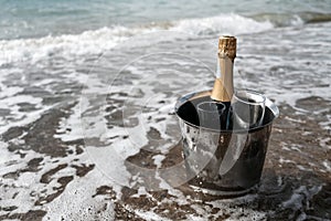 Ice bucket with champagne bottle and glasses on a beach surrounded by sea. Ocean waves chilling drinks, luxury vacation for two.