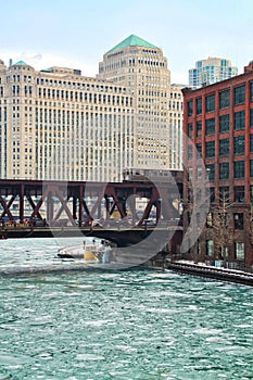 An ice chunk covered, frozen Chicago River is aqua blue and runs through Chicago Loop while el train passes over water