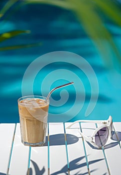 Ice coffee Cyprus Frappe Fredo against blue clear water of the swimming pool, on white table, with sunglasses . Summer
