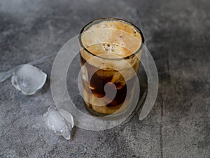Ice coffee in a tall glass, ice cubes on a gray wooden table. Cold summer drink with tubes on a black background with