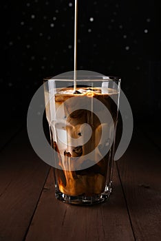 Ice coffee on a wood table with cream pouring into glass.