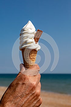 An ice cream cone with chocolate flake being held against a blue sky at the beach, with a shallow depth of field