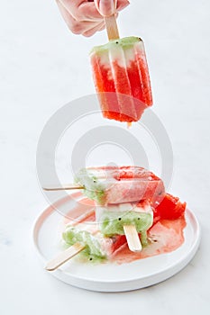 Ice cream lolly in a plate with splashes. A woman`s hand holds a melting ice cream on a white marble background with