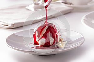 Ice cream scoop with pouring berry sauce from spoon on white plate close-up