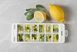 Ice cube tray with mint, lemon and water on grey, top view