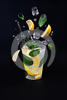 Ice cubes, slices of lemon and mint leaves splashing into glass of lemonade on black