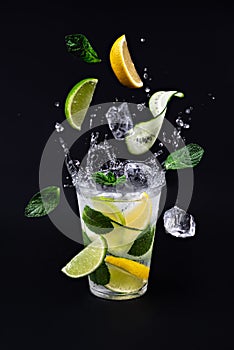 Ice cubes, slices of lime. lemon, cucumber and mint leaves splashing into glass of lemonade on black