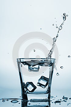 Ice cubes splashing into glass, ice cube dropped into glass of water, fresh, cold water, isolated on blue, caribian blue backgrou photo