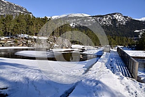 Ice on the river at Valle Norway, Feb 2023. Snow on the ground and mountains behind. Bridge over a waterfall.