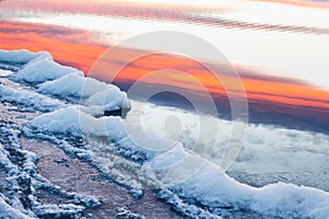 Ice on the shore of lake. Sky with pink clouds reflected in the cold water at sunset