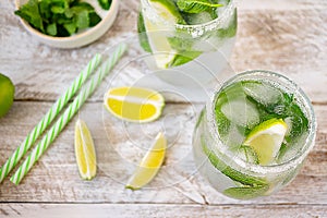 Ice summer lemonade with lime and mint in glasses stands on a light wooden table. Near lay striped cocktail tubes and three slices