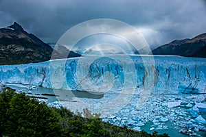 Ice wall of a glacier with some pieces of ice in the water in front of a forest