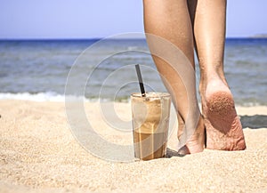 Iced coffee and female legs on a sandy beach