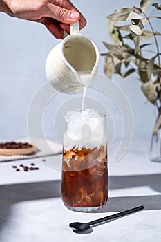 Iced coffee latte in a tall glass with a hand pouring milk from a milk jug on a blue background with coffee beans, branch and