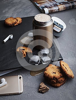 Iced coffee in a steel thermo mug, with almond milk, ice cubes and homemade cookies on a black stone background.
