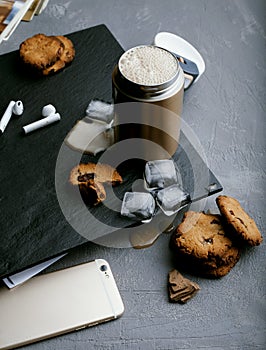 Iced coffee in a steel thermo mug, with almond milk, ice cubes and homemade cookies on a black stone background. Cold summer drink