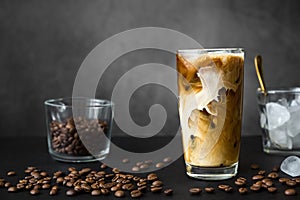 Iced coffee in tall glass with cream, container with ice and spoon, cocktail straw and coffee beans on dark background with copy