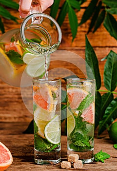 Iced summer drink with citrus fruits and mint pouring from transparent jug into glass