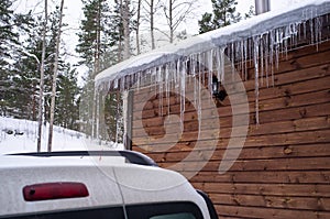 Icicles hang from the roof of a log house near which a car is carelessly parked, which can be damaged by falling ice