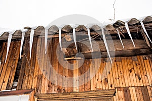 Icicles hang from the roof of a wooden house in the countryside, on a frosty cloudy day