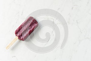 An icy purple grape popsicle rests at an angle on a white marble countertop with copy space; sweet dessert
