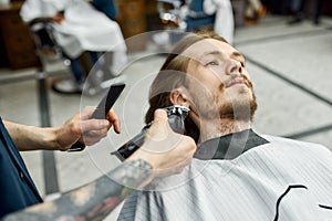Ideal beard shape. Barber with tattoo on his arm holding hair clipper and trimming beard of his client. Barbershop
