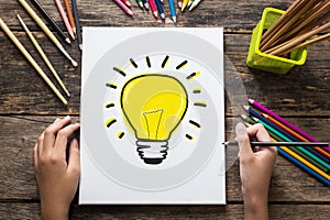 Ideas Concept , Hand of girl drawing a light bulb on paper and colorful pencils on wooden table