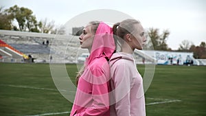 Identical twin sisters in sportswear stand outdoor at stadium field back to back