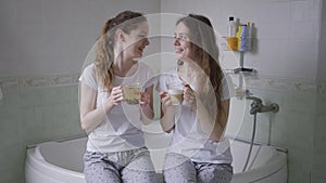 Identical twin sisters talking sitting on bathtub with tea and coffee in cups. Smiling positive young Caucasian