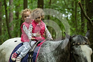 Identical twins enjoying horseback riding in the woods, young pretty girls with blond curly hair on a horse with backlit leaves