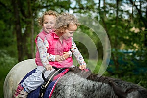 Identical twins enjoying horseback riding in the woods, young pretty girls with blond curly hair on a horse with backlit leaves