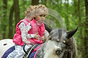 Identical twins enjoying horseback riding in the woods, young pretty girls with blond curly hair on a horse with backlit leaves