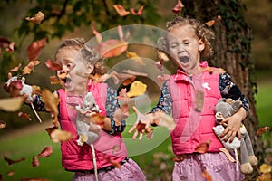 Identical twins having fun with autumn leaves, blond cute curly girls, happy family, beautiful girls in pink jackets