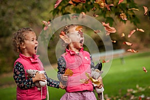 Identical twins having fun with autumn leaves, blond cute curly girls, happy family, beautiful girls in pink jackets