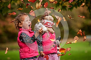 Identical twins having fun with autumn leaves, blond cute curly girls, happy family, beautiful girls in pink jackets